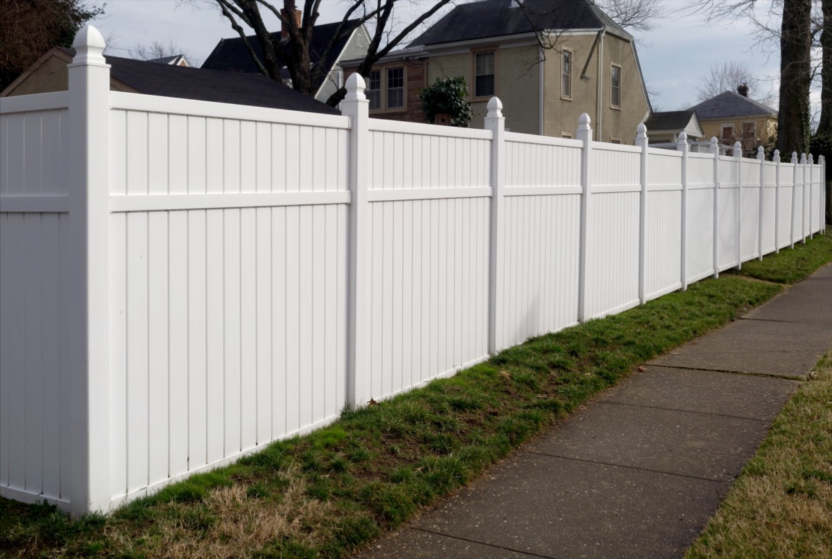 A white vinyl privacy fence with decorative post caps running along a grassy strip beside a sidewalk
